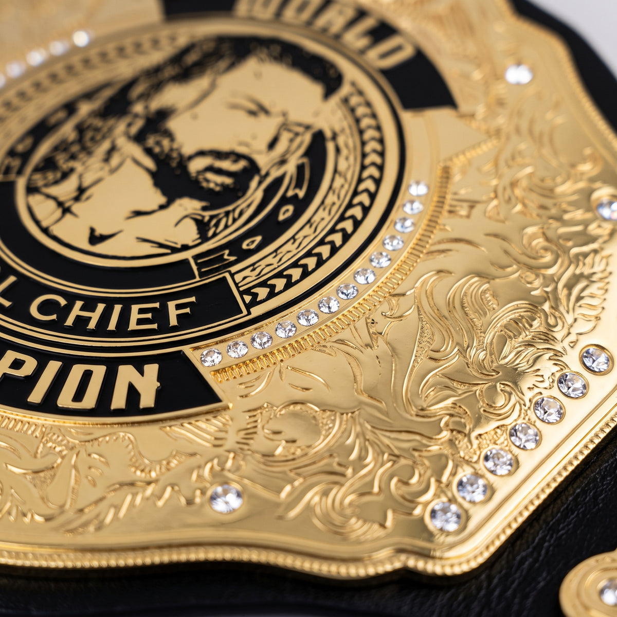 Close-up of a gold championship belt with engraved details, black lettering, and clear rhinestones on a black surface.