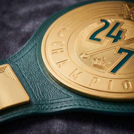 Close-up of a championship belt with a textured dark green leather strap and a large gold-toned medallion featuring the number 24 and the word CHAMPION.