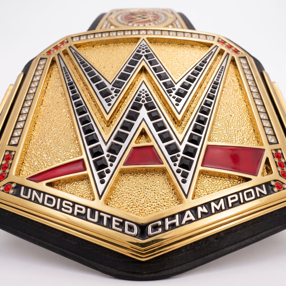 Close-up of a WWE Undisputed Champion belt featuring a large gold-tone central plate with a black and silver WWE logo, red accents, and the words 'UNDISPUTED CHAMPION' on a white background.