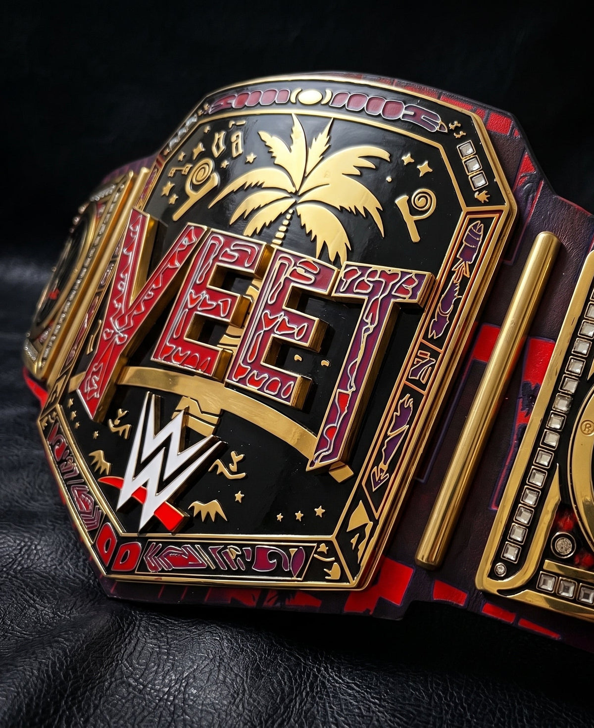 A close-up of a WWE 'Vet' championship belt with ornate gold and red detailing, black enamel, and a palm tree motif, photographed on a dark textured surface.