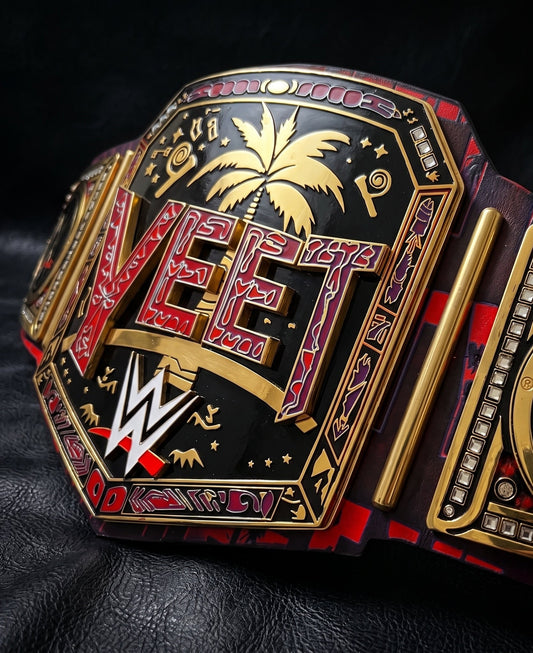 A close-up of a WWE 'Vet' championship belt with ornate gold and red detailing, black enamel, and a palm tree motif, photographed on a dark textured surface.