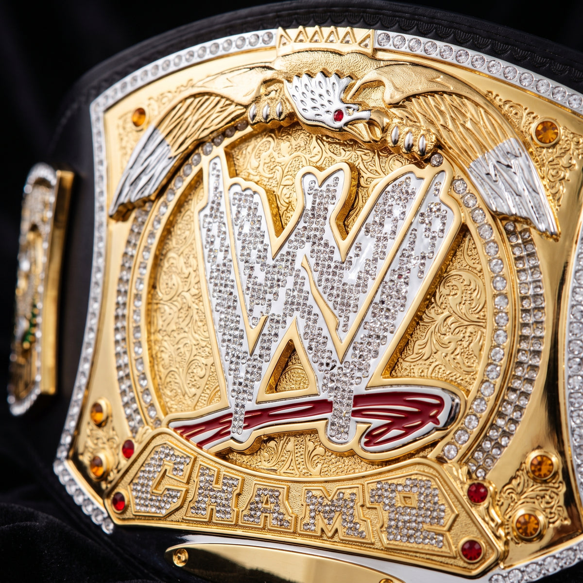 Close-up of a WWE championship belt plate with a large gold and silver eagle design, rhinestone-embellished WWE logo, and the word CHAMP below on a black velvet background.