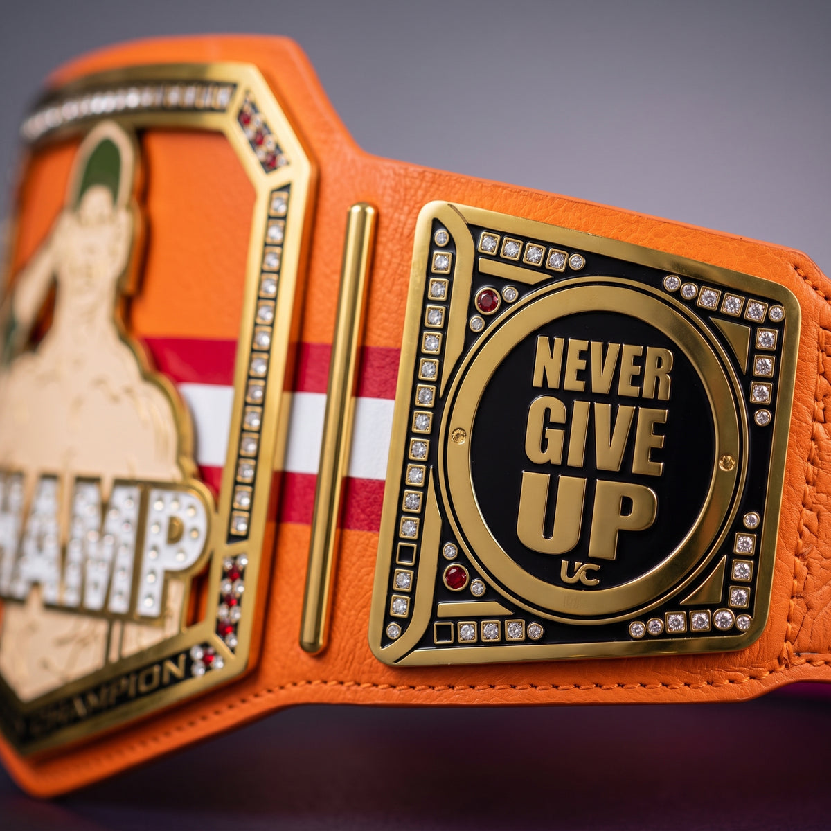 Close-up of an orange championship belt with gold-tone metal plates, black center panels, rhinestone accents, and the words "NEVER GIVE UP UC" on one side.