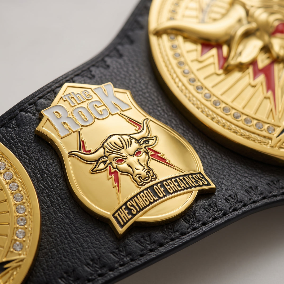 Close-up of a black leather belt featuring a gold-tone shield-shaped metal emblem with a bull head and the words 'The Rock' and 'The Symbol of Greatness'.