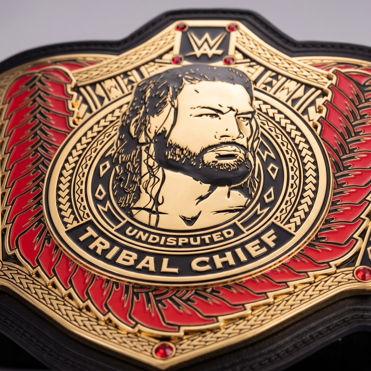 Close-up of a WWE Tribal Chief Undisputed Championship belt featuring a gold-toned central medallion with a portrait, red feather-like accents, and black border details.
