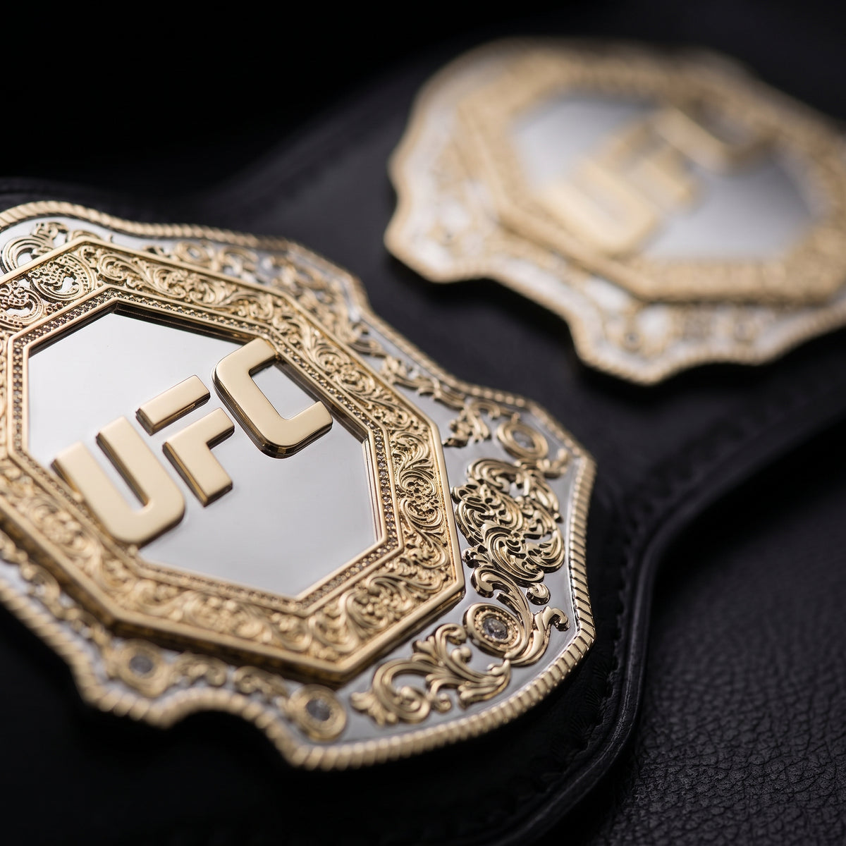 Close-up of a black leather UFC belt with ornate gold-tone metal plates and a visible UFC logo, photographed against a dark background.