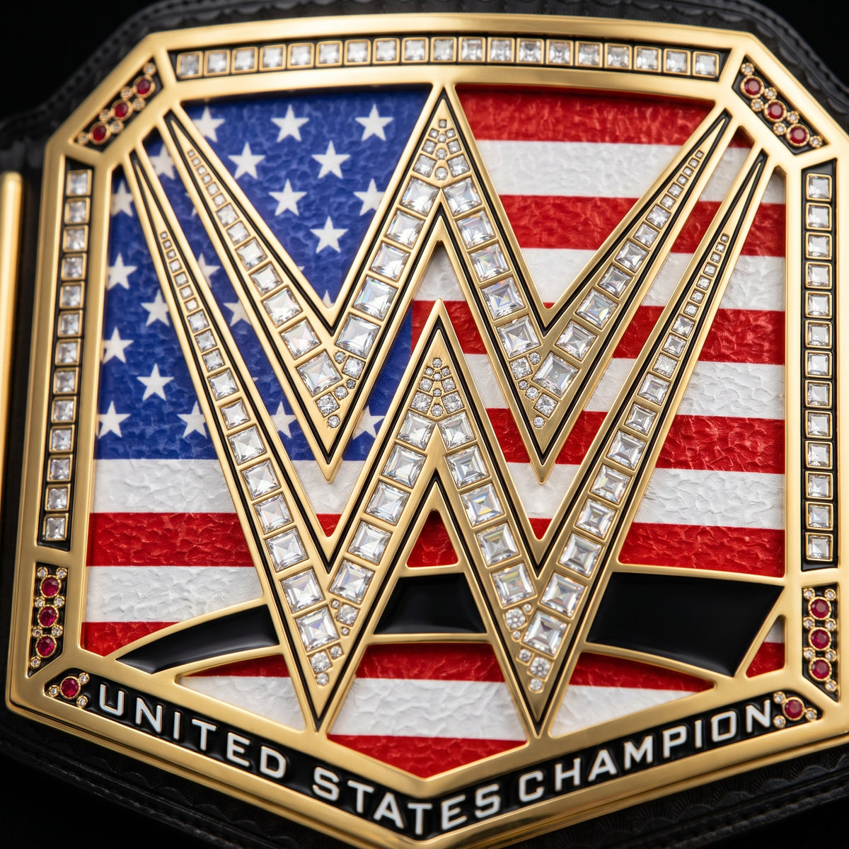 Close-up of a championship belt plate featuring a large interlocking WWE-style 'W' emblem over an American flag background, with gold-tone metal, clear stones, and the text 'UNITED STATES CHAMPION' along the bottom.