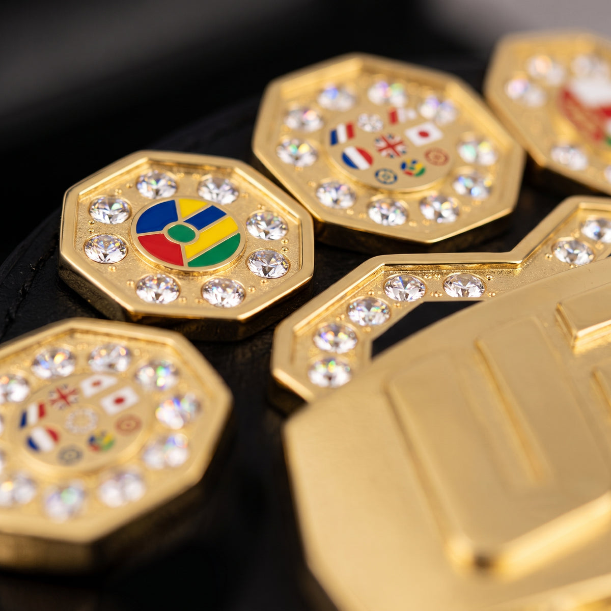 Gold-toned octagonal belt buckles adorned with clear rhinestones, including one featuring a colorful round emblem and others with small national flag motifs, shown on a black background.
