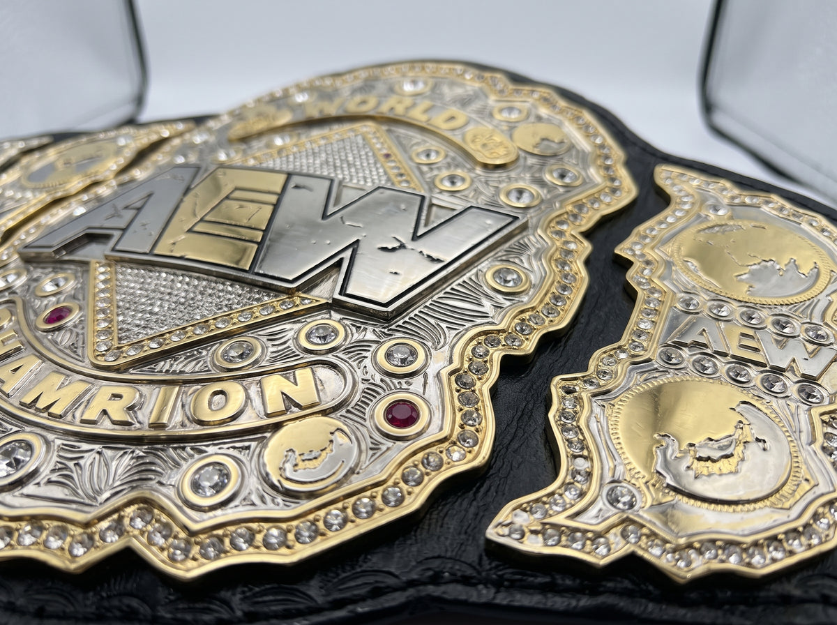 Close-up of an ornate AEW Championship belt with gold and silver metal plates, rhinestone accents, and a black strap, shown inside a clear display case.