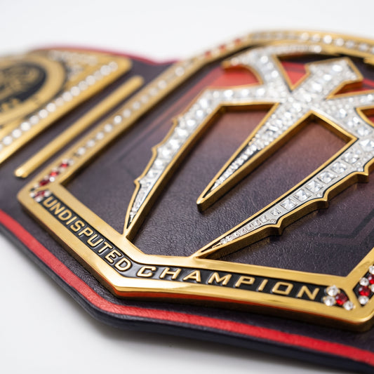 Close-up of a championship belt plate with gold metal detailing, clear rhinestones, and a dark red and black leather strap on a white background.