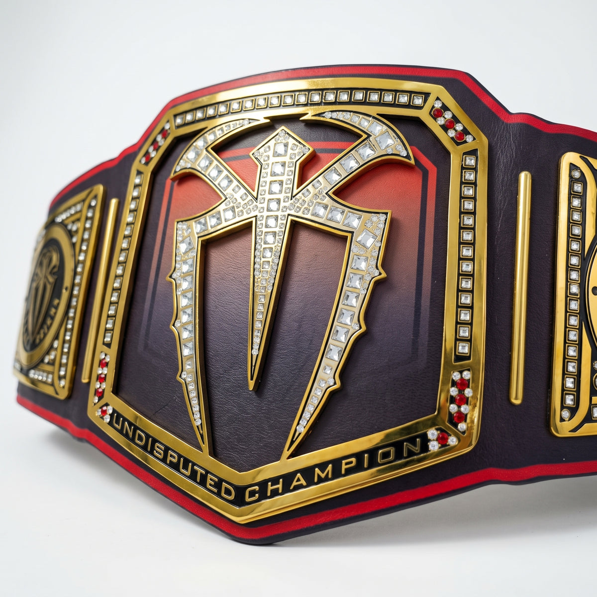 A close-up of a championship belt featuring a large central plate with a stylized emblem outlined in clear rhinestones, gold trim, and a red-to-black gradient background. The words 'UNDISPUTED CHAMPION' are visible along the lower edge.