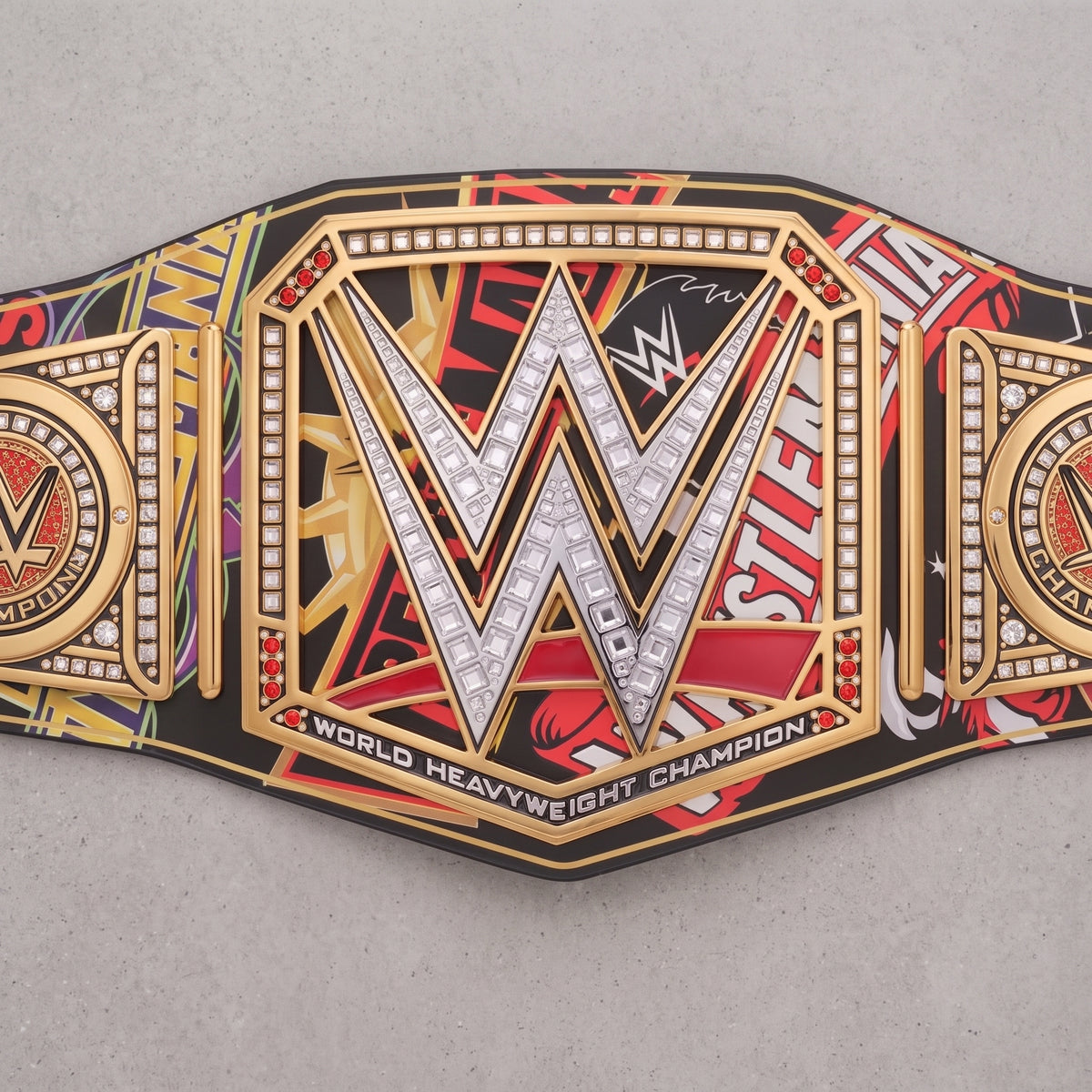 A WWE World Heavyweight Championship belt laid flat on a light gray surface. The belt features a large central gold and silver 'W' with rhinestone-like detailing, flanked by red, black, and gold graphic panels with WWE branding and text.