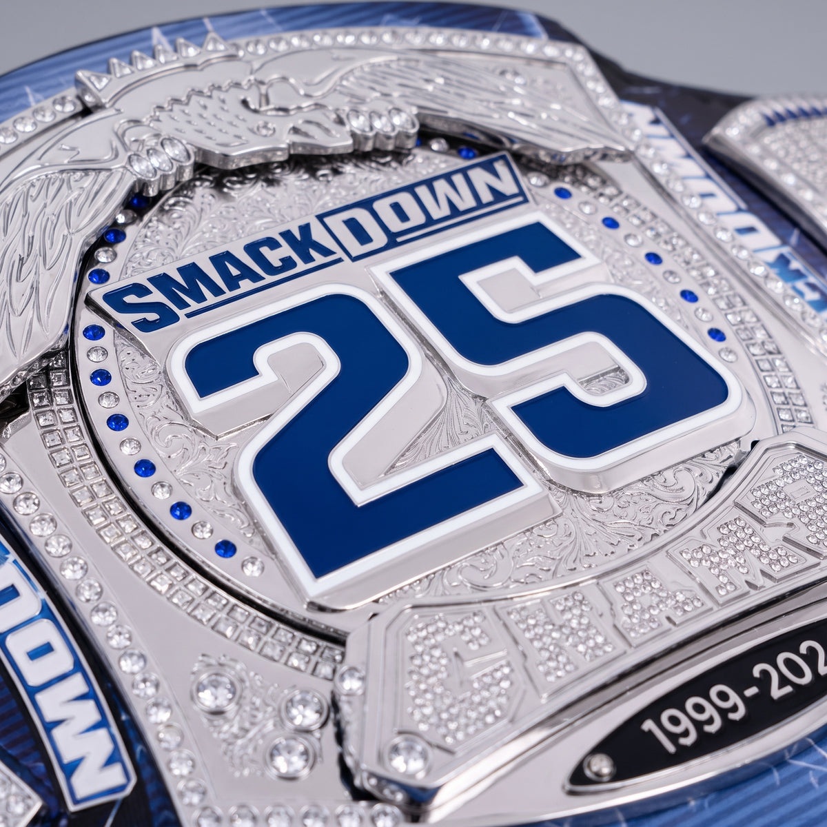 A close-up of a custom championship belt featuring a silver-toned plate with blue and white details, rhinestones, and the text 'SMACKDOWN 25' and '1999-2025'.