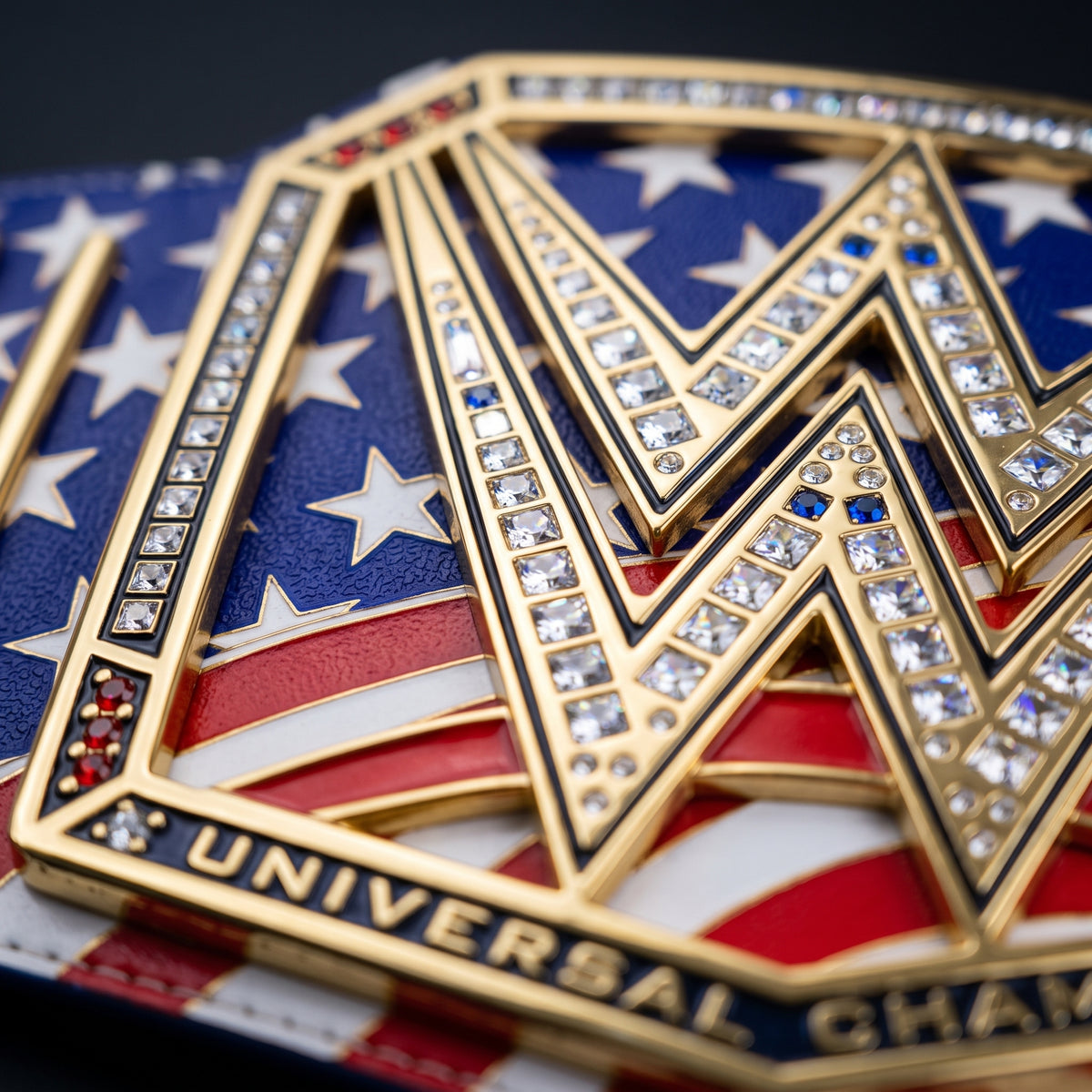 Close-up of a gold Universal Championship belt buckle featuring an American flag design with blue, red, and white enamel, set with clear rhinestones and small blue accents.