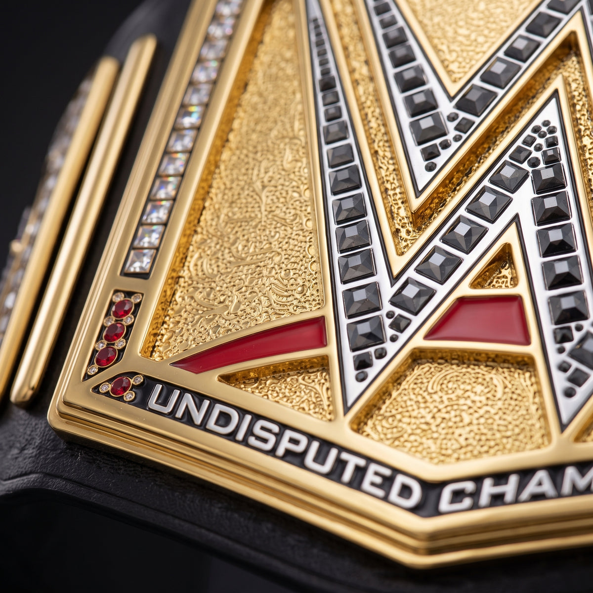 Close-up of a gold championship belt plate with black and silver geometric details, red accents, and the text 'UNDISPUTED CHAMPION' on a dark background.