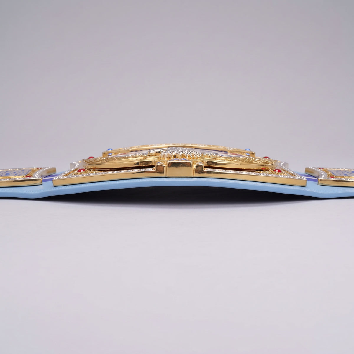 A close-up of a light blue belt featuring an ornate gold central buckle embellished with clear crystals and small red and blue gemstones, set against a neutral gray background.