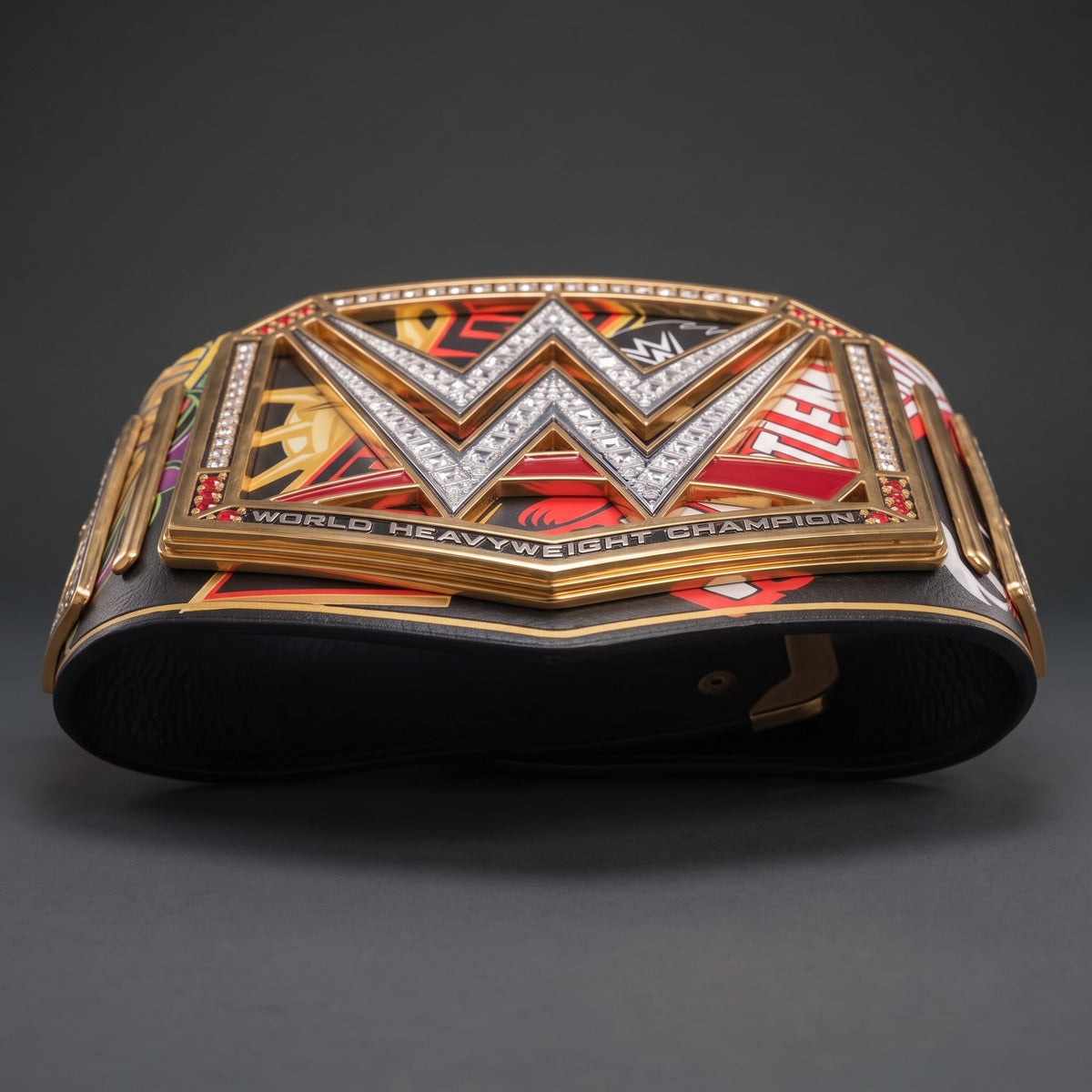 A replica WWE World Heavyweight Championship belt displayed on a dark gray background. The belt features a large gold-toned central plate with a silver crystal-embellished 'W' logo, flanked by red and black panels with various championship graphics and text.