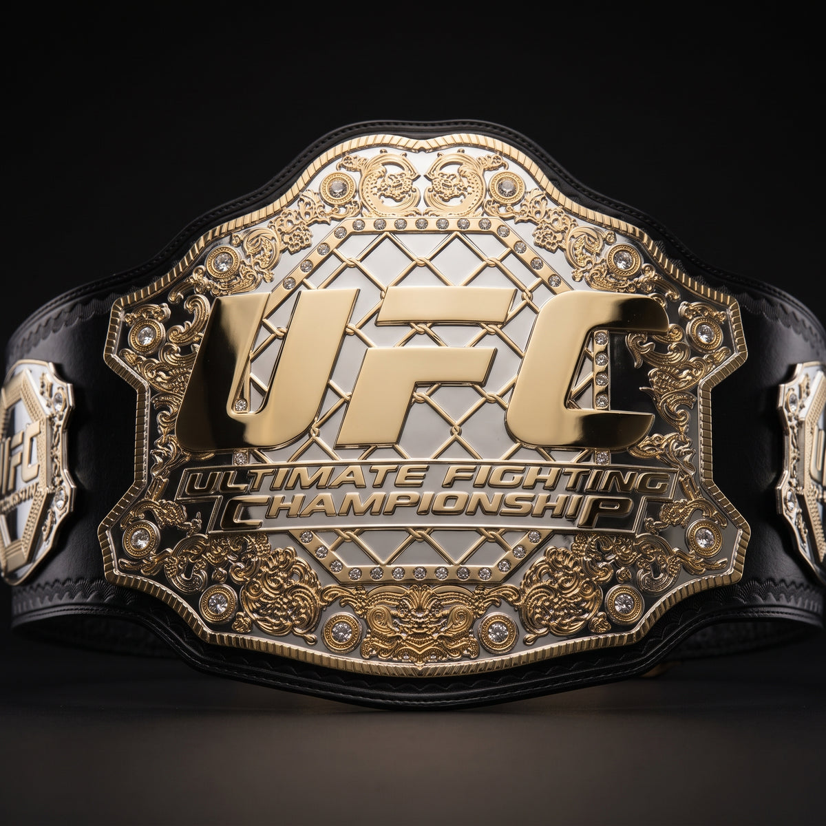 A close-up studio shot of a UFC Ultimate Fighting Championship belt, showing the ornate gold and silver central plate with the large gold UFC logo, black strap sections, and decorative embossed details on a dark background.
