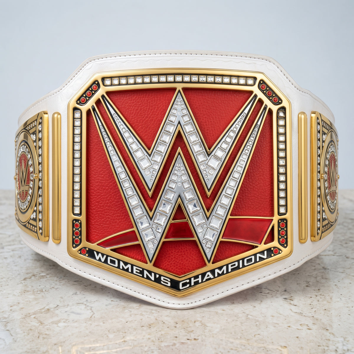 A WWE Women's Champion belt displayed on a light marble surface against a light gray background. The belt features a large red central panel with a silver 'W' outlined in gold, flanked by gold side plates with red and black details, and a white leather strap.